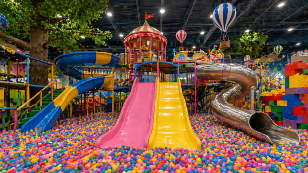 A vibrant indoor playground filled with colorful slides and a ball pit. Dominating the foreground is a large ball pit overflowing with thousands of multi-colored plastic balls in shades of red, blue, yellow, green, pink, and purple. Two large, brightly colored slides, one pink and one yellow, descend from a high platform and empty into the ball pit. To the left, a blue slide with yellow diagonal stripes snakes downwards, and green stairs with colorful railings lead upwards. In the background, a large, silver tubular slide curves into the frame from the right. The environment is a playful and energetic space designed for children, with artificial trees, decorative hot air balloons, and stacked colorful building blocks visible. The lighting is bright and even, illuminating the cheerful atmosphere. The composition uses a wide-angle perspective to capture the expansive nature of the playground, with the slides and ball pit occupying most of the frame. The overall style is playful and somewhat fantastical, evoking a sense of fun and excitement.